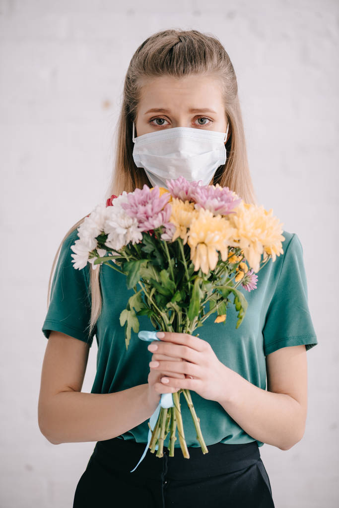 blonde woman in medical mask holding flowers in hands and looking at camera  - Photo, Image