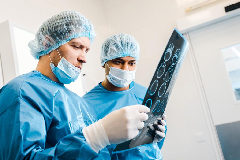 gut aussehende Ärzte in Uniformen und medizinischen Masken, die in der Klinik das Röntgenbild betrachten  - Foto, Bild