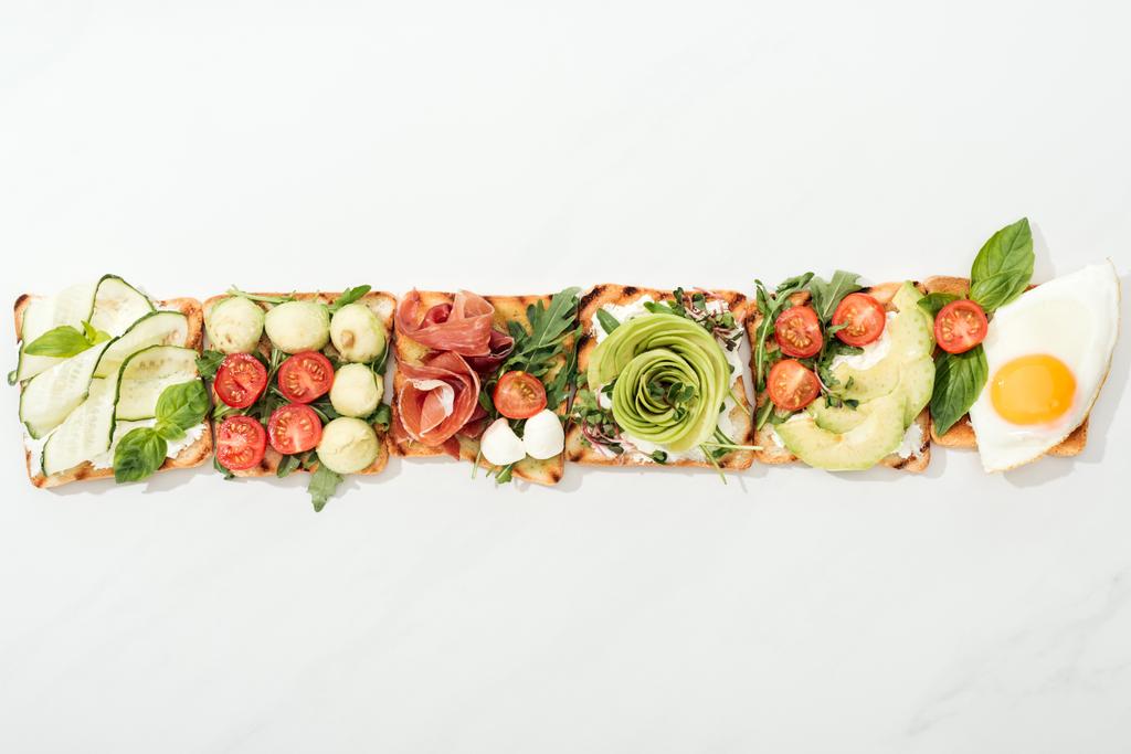 Вид сверху на тосты с нарезанными овощами, жареным яйцом и prosciutto на белой поверхности
 - Фото, изображение