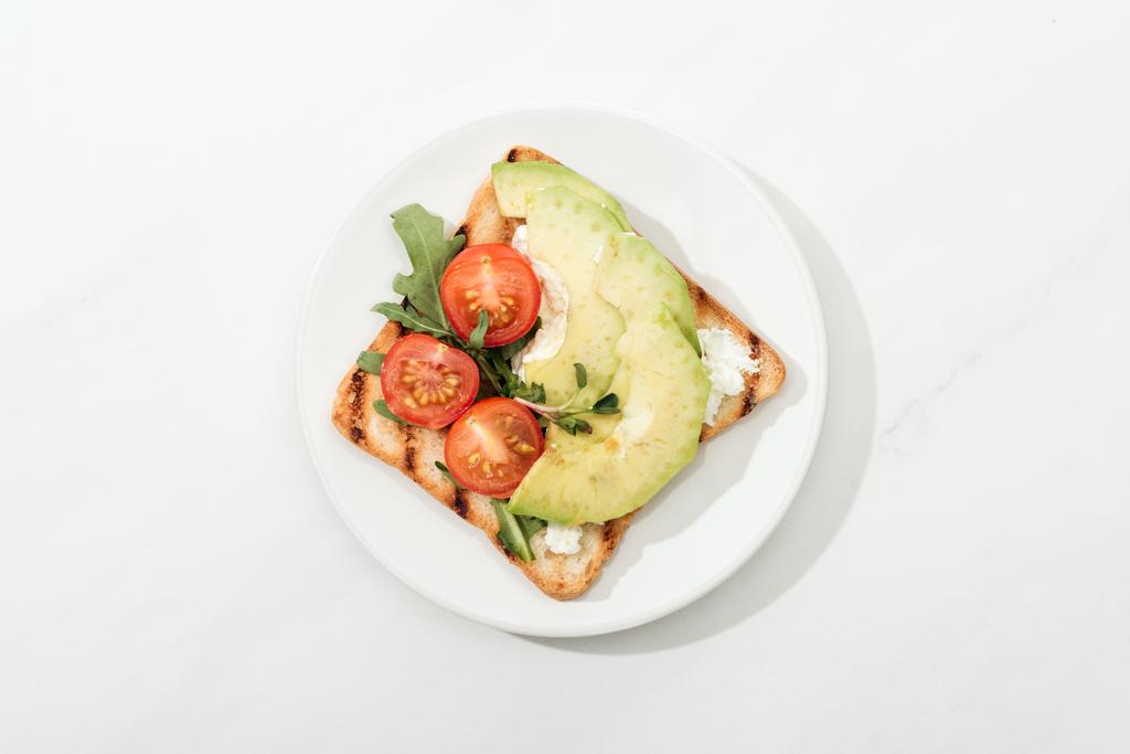 Vista dall'alto di pane tostato con pomodorini ciliegia e avocado su piatto su superficie bianca
 - Foto, immagini