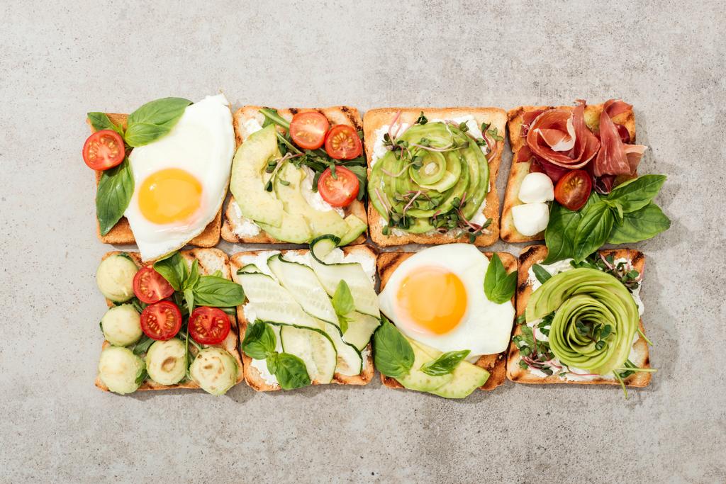 Vista superior de tostadas con verduras, huevos fritos y jamón en superficie texturizada
 - Foto, Imagen