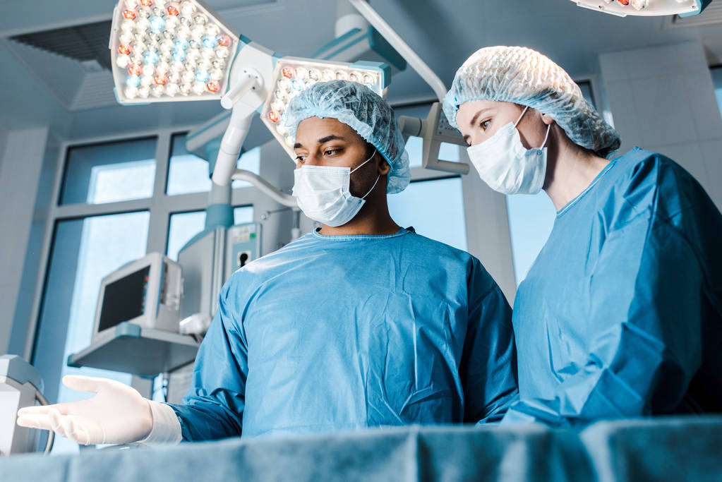  Krankenschwester in Uniform und Maske und Arzt zeigt mit den Fingern im Operationssaal  - Foto, Bild