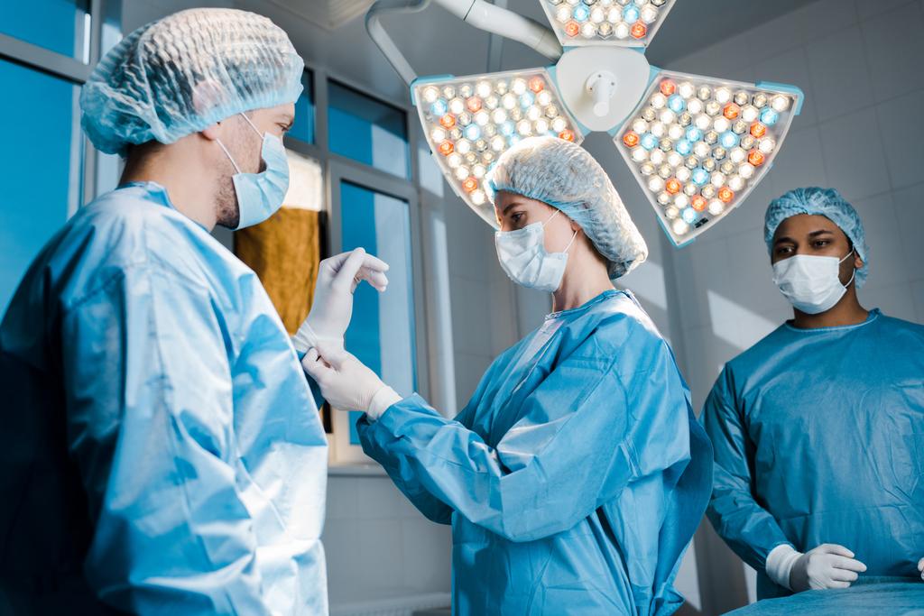 Krankenschwester in Uniform und medizinischer Maske legt Latex-Handschuh auf die Hand des Arztes  - Foto, Bild