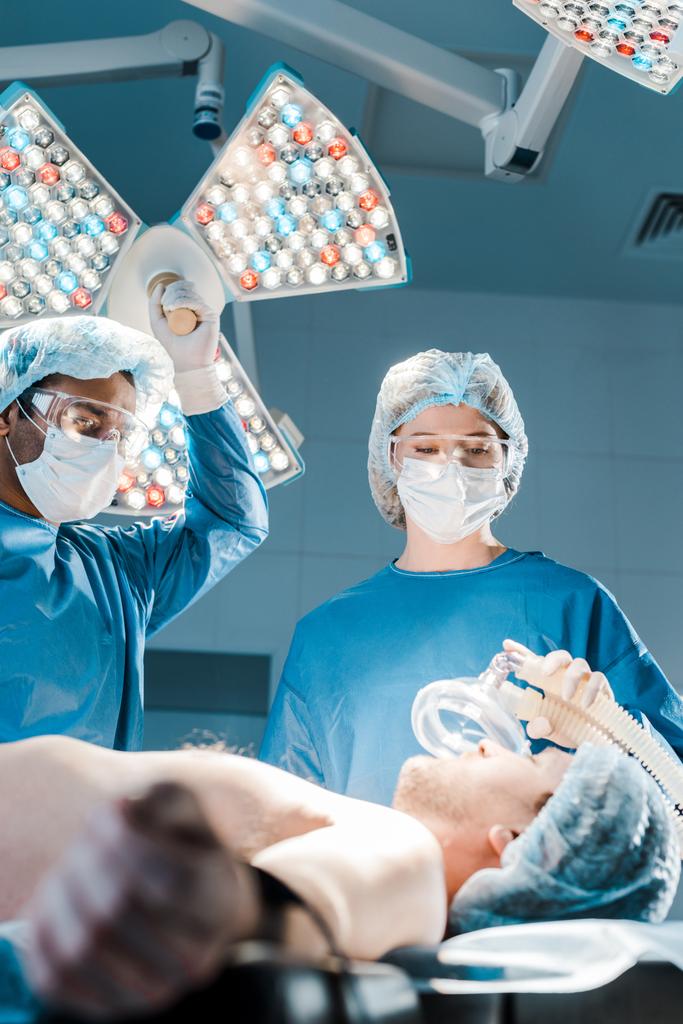 selective focus of doctor and nurse in uniforms and medical caps putting mask on patient  - Photo, Image