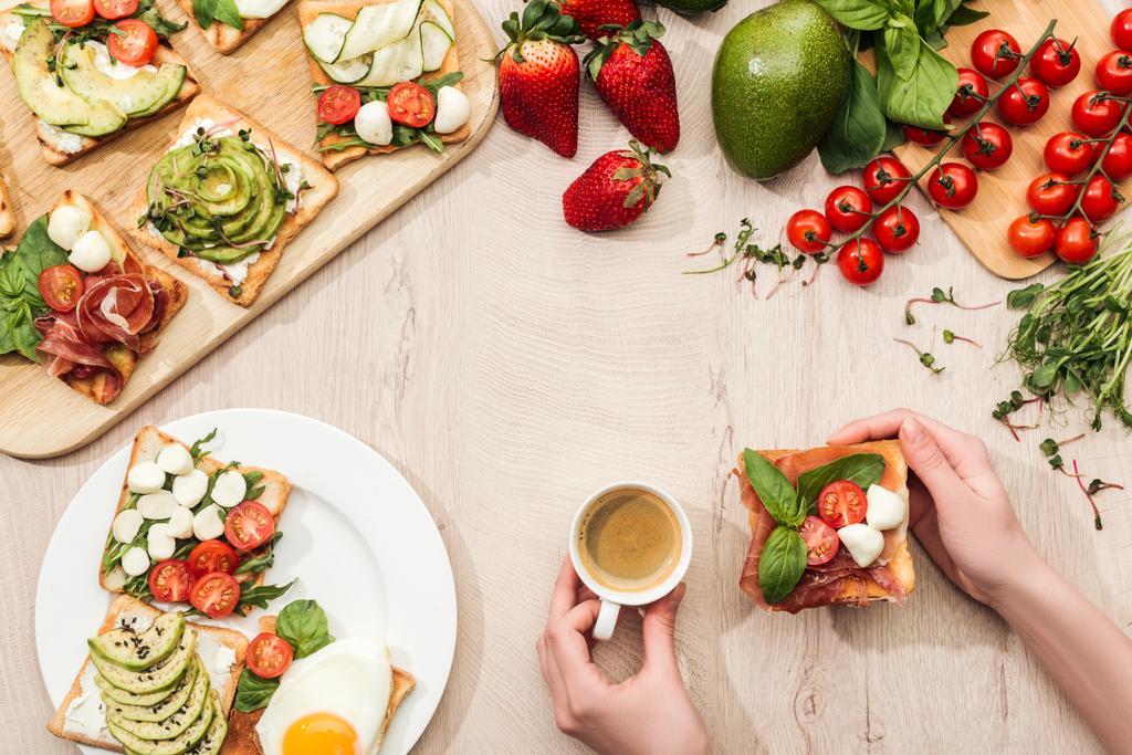 食卓の食材、緑の野菜と生ハムのトーストとコーヒーのカップを保持している女性のトップ ビュー - 写真・画像