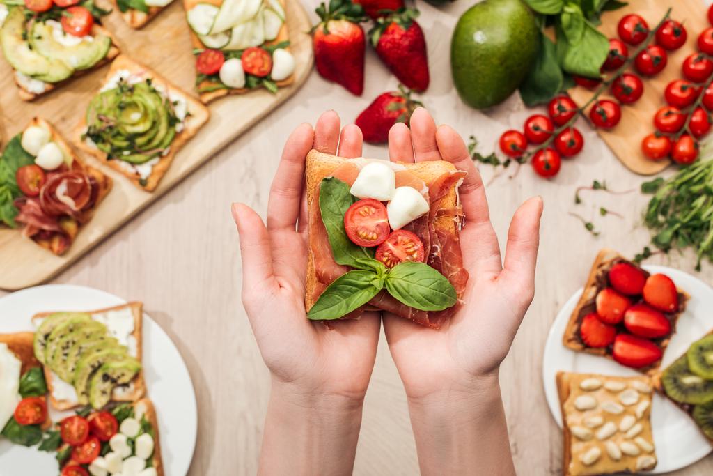selective focus of woman holding toast with cherry tomatoes and prosciutto over wooden table with greenery and ingredients - Photo, Image