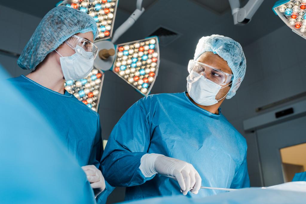 Krankenschwester in Uniform und Chirurg mit Mütze schauen sich im Operationssaal an  - Foto, Bild