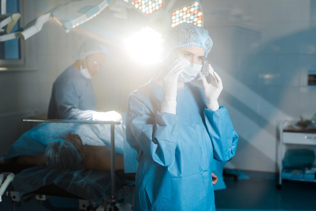 foco seletivo da enfermeira em uniforme e máscara médica adiando óculos na sala de cirurgia
 - Foto, Imagem