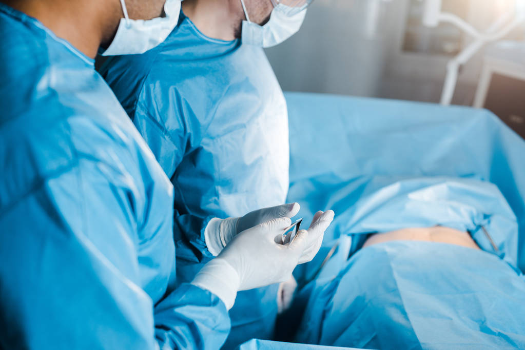 κομμένη θέα της νοσοκόμας σε στολή και ιατρική μάσκα δίνοντας ιατρικό εξοπλισμό σε χειρουργό  - Φωτογραφία, εικόνα