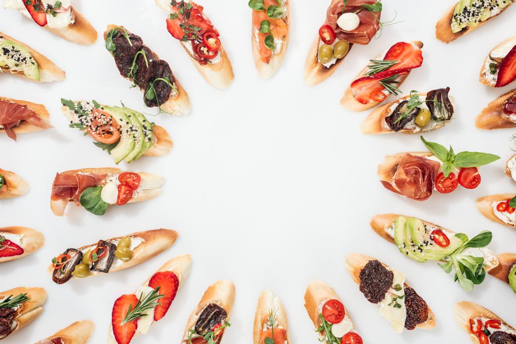 top view of round frame made of italian bruschetta with salmon, prosciutto, herbs and various fruits with vegetables on white - Photo, Image