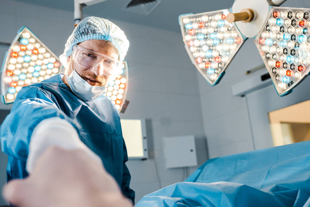 selective focus of surgeon in uniform and medical mask looking at camera and holding hand of patient  - Photo, Image