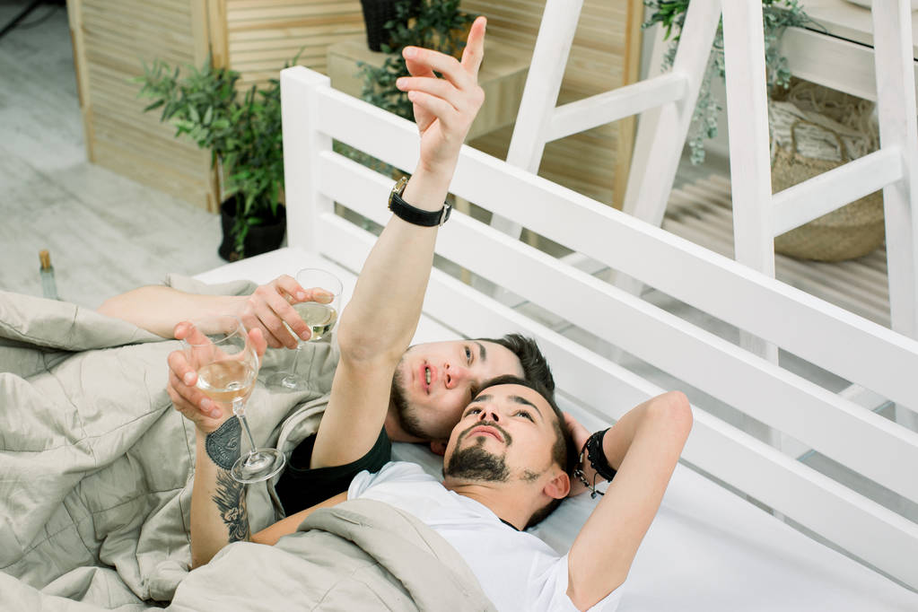 Два симпатичных парня пьют белое вино. валялся дома в постели. Молодая пара геев лежит под одеялом на кровати в светлой комнате
 - Фото, изображение