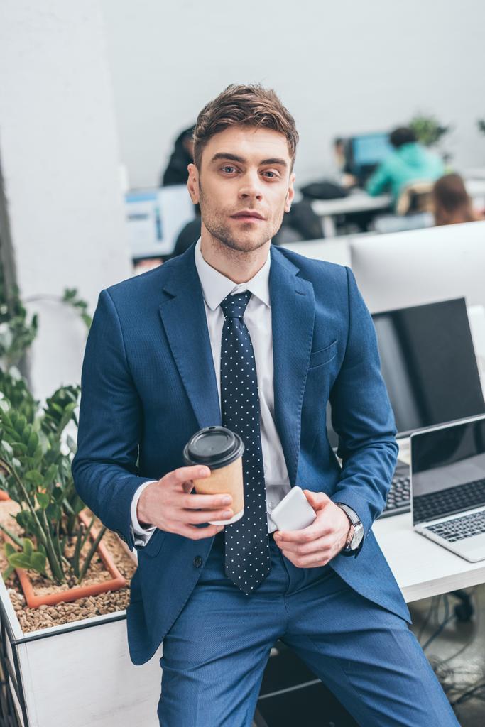 confident businessman holding smartphone and paper cup and looking at camera - Photo, Image
