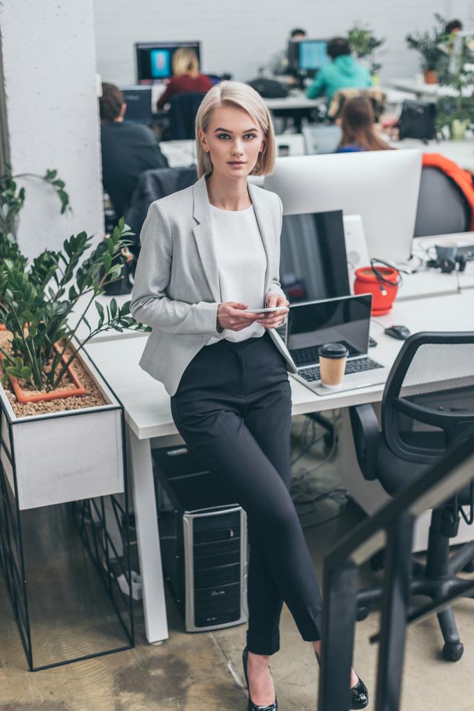 pretty blonde businesswoman in formal wear standing by workplace and looking at camera - Photo, Image