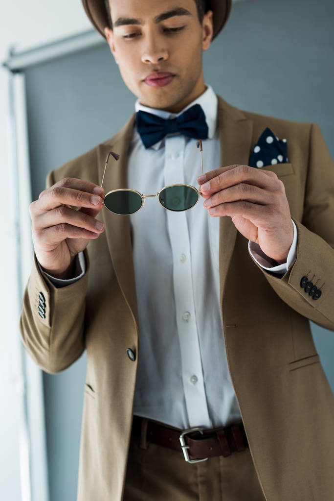 homem de raça mista elegante em terno e gravata arco segurando óculos de sol em cinza
 - Foto, Imagem