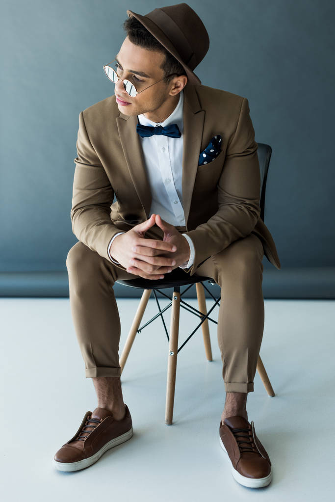 elegante uomo di razza mista in giacca e cravatta e occhiali da sole seduto sulla sedia e in posa su grigio e bianco
 - Foto, immagini