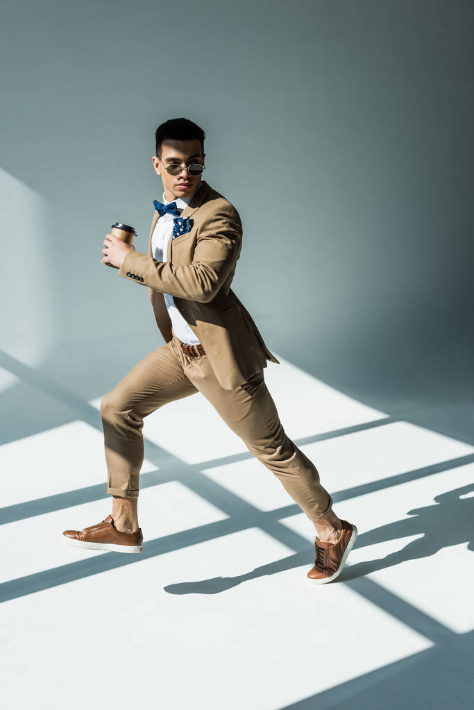 stijlvolle mixed race man in pak houden van koffie te gaan en springen op grijs met zonlicht - Foto, afbeelding