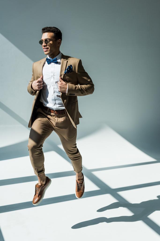 stylischer Mixed Race Mann in Anzug und Sonnenbrille springt auf grau mit Sonnenlicht - Foto, Bild