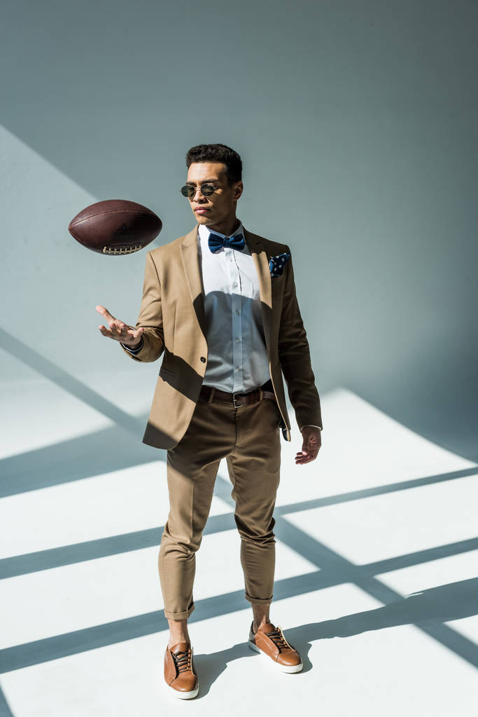 stylischer Mischlingsmann im Anzug, der Fußballball auf grau mit Sonnenlicht wirft - Foto, Bild