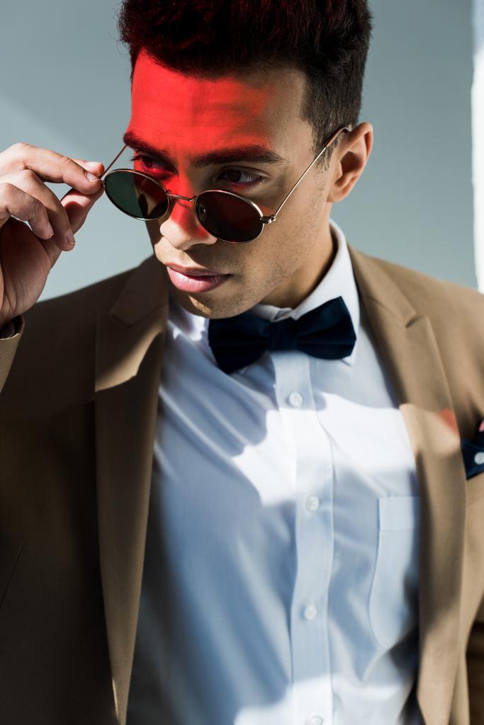 стильный смешанный гонщик в костюме и солнцезащитных очках, позирующий на сером с красным светом
 - Фото, изображение