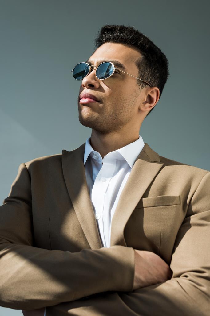 elegante uomo di razza mista in tuta e occhiali da sole in posa con braccia incrociate su grigio con luce solare
 - Foto, immagini