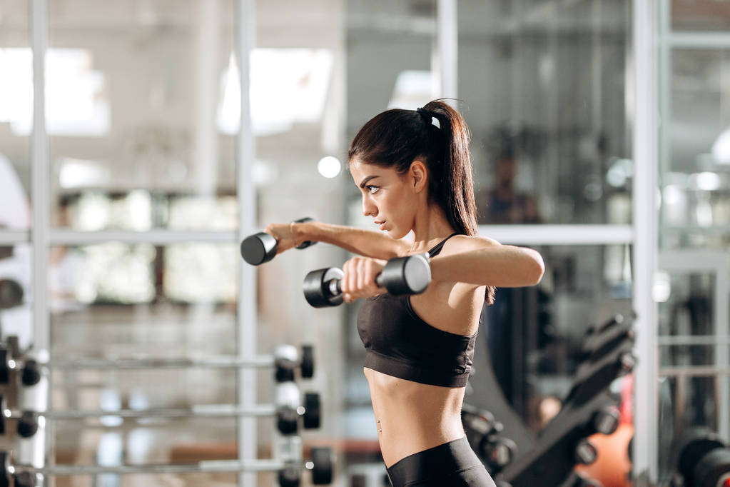 Hermosa chica atlética vestida con top deportivo negro y medias acumula músculos con mancuernas en el gimnasio
 - Foto, imagen