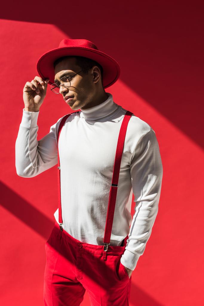 fashionable mixed race man in hat and suspenders holding glasses while posing on red - Photo, Image