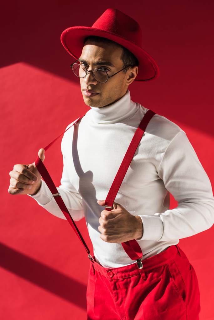 homem de raça mista na moda em chapéu e suspensórios olhando para a câmera enquanto posando no vermelho
 - Foto, Imagem