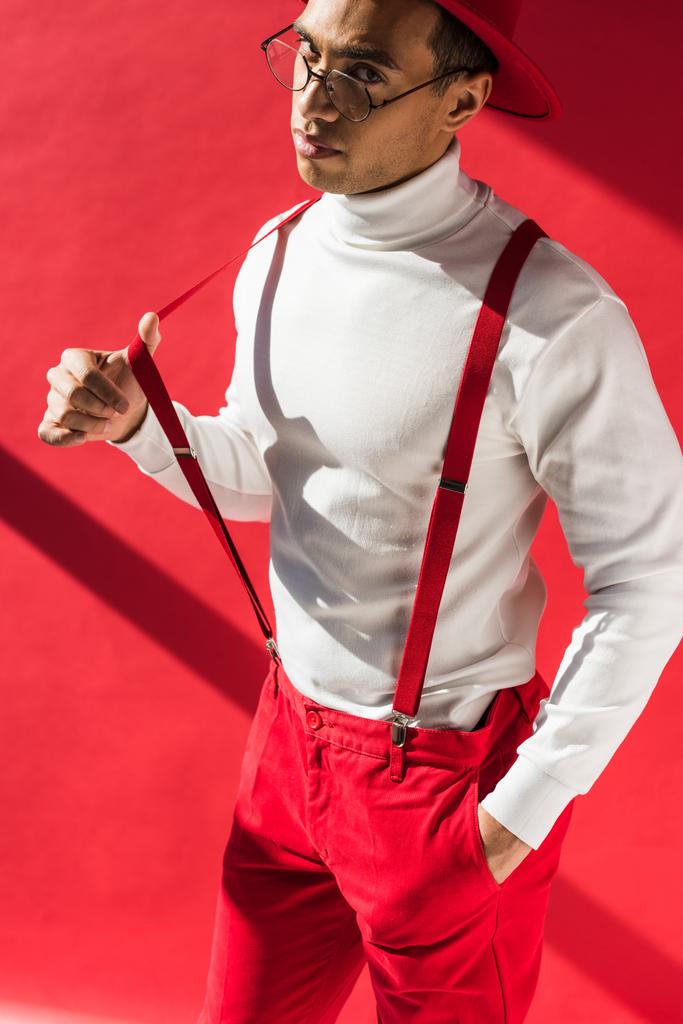 fashionable mixed race man in hat and suspenders looking at camera while posing on red - Photo, Image