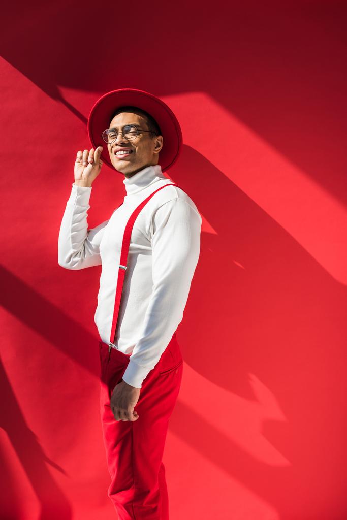 hombre de raza mixta de moda en sombrero y tirantes sonriendo y posando en rojo con espacio de copia
 - Foto, Imagen