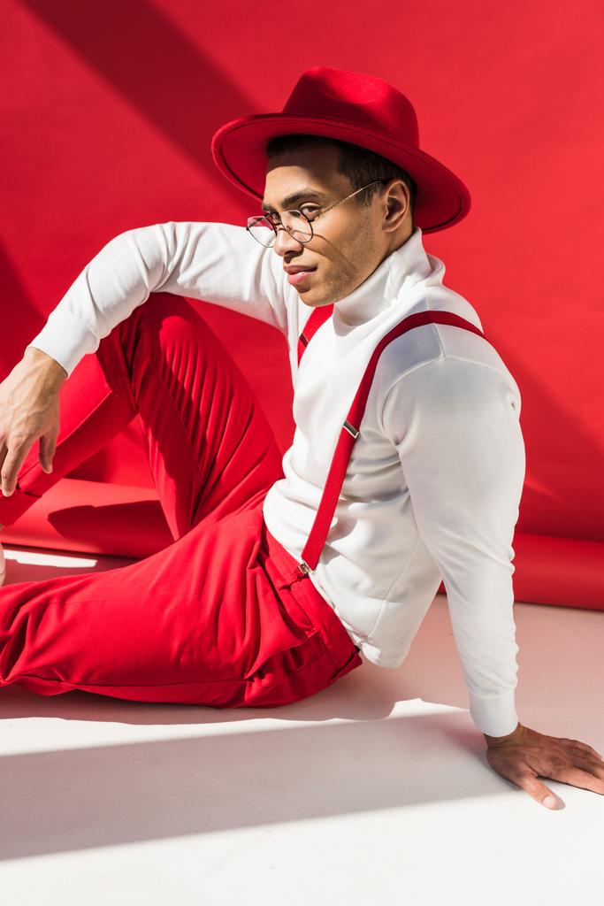 modieuze mixed race man in Hat en bretels zittend en poseren op rood - Foto, afbeelding