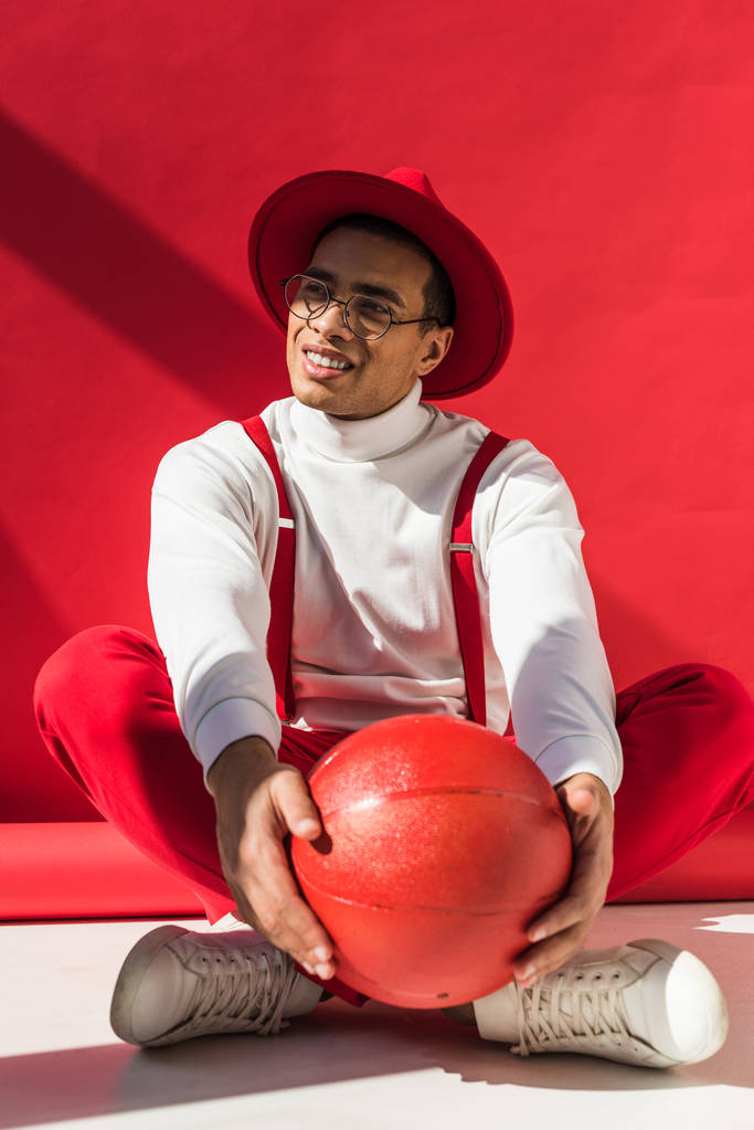 stylischer Mixed Race Mann mit Hut und Hosenträgern sitzend, lächelnd und posierend mit Basketball auf Rot - Foto, Bild