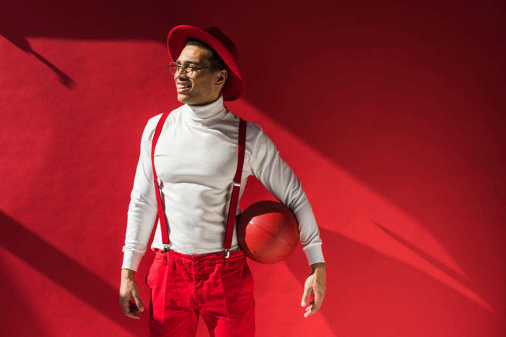 κομψό ανάμεικτο άντρας με καπέλο και τιράντες που ποζάρει με μπάσκετ στο κόκκινο με χώρο αντιγραφής - Φωτογραφία, εικόνα