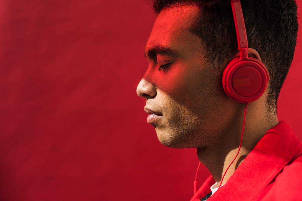 vue latérale du beau mixte homme dans les écouteurs avec les yeux fermés isolé sur rouge avec espace de copie
 - Photo, image
