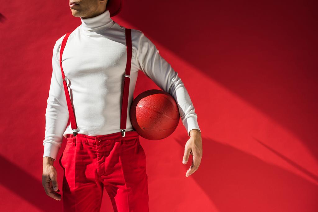 обрезанный вид стильного смешанного расового человека в подтяжках позирует с баскетболом на красном с копировальным пространством
 - Фото, изображение
