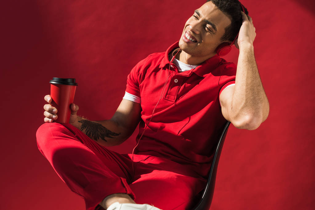 Стильный мужчина смешанной расы в наушниках сидит, держит кофе, чтобы пойти и улыбается на красный
 - Фото, изображение