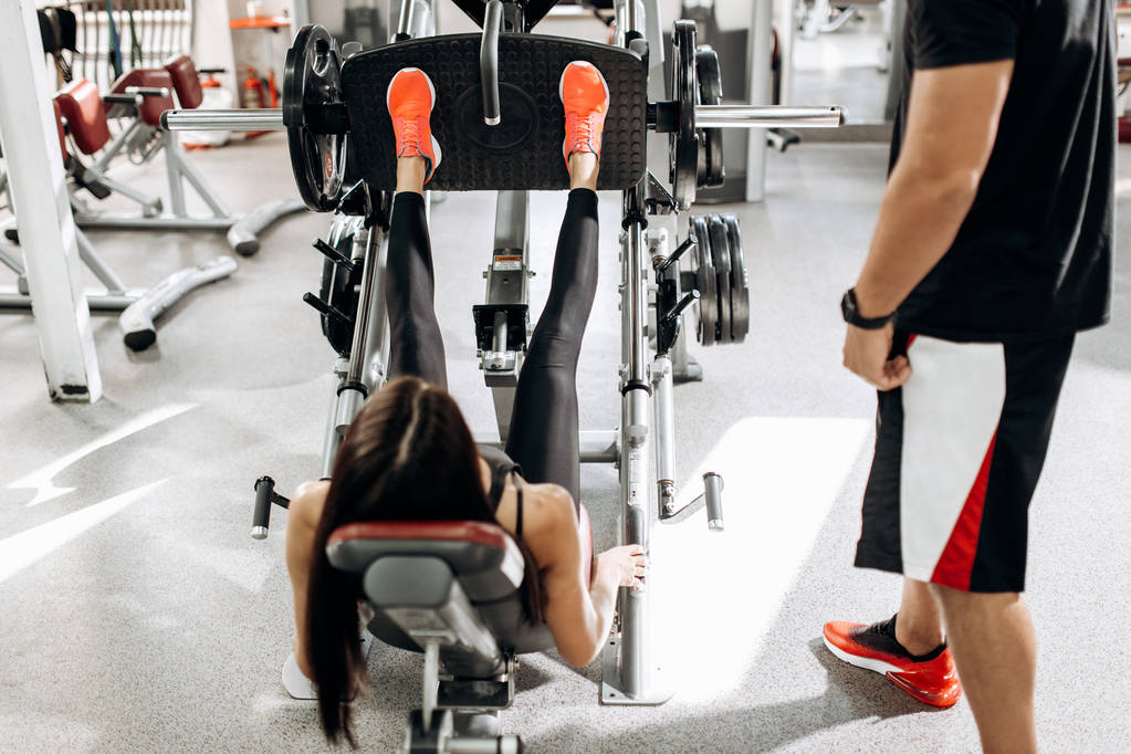 Atletik kız siyah spor kıyafetlerini giymiş ile bacaklarını bir antrenör gözetiminde spor salonunda ağırlık kaldırma - Fotoğraf, Görsel