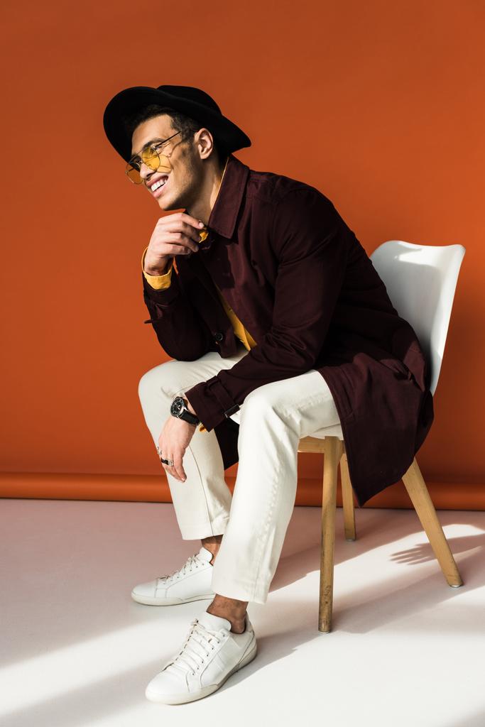 stijlvolle mixed race man in Hat en zonnebrillen zittend op de stoel en glimlachend op oranje - Foto, afbeelding