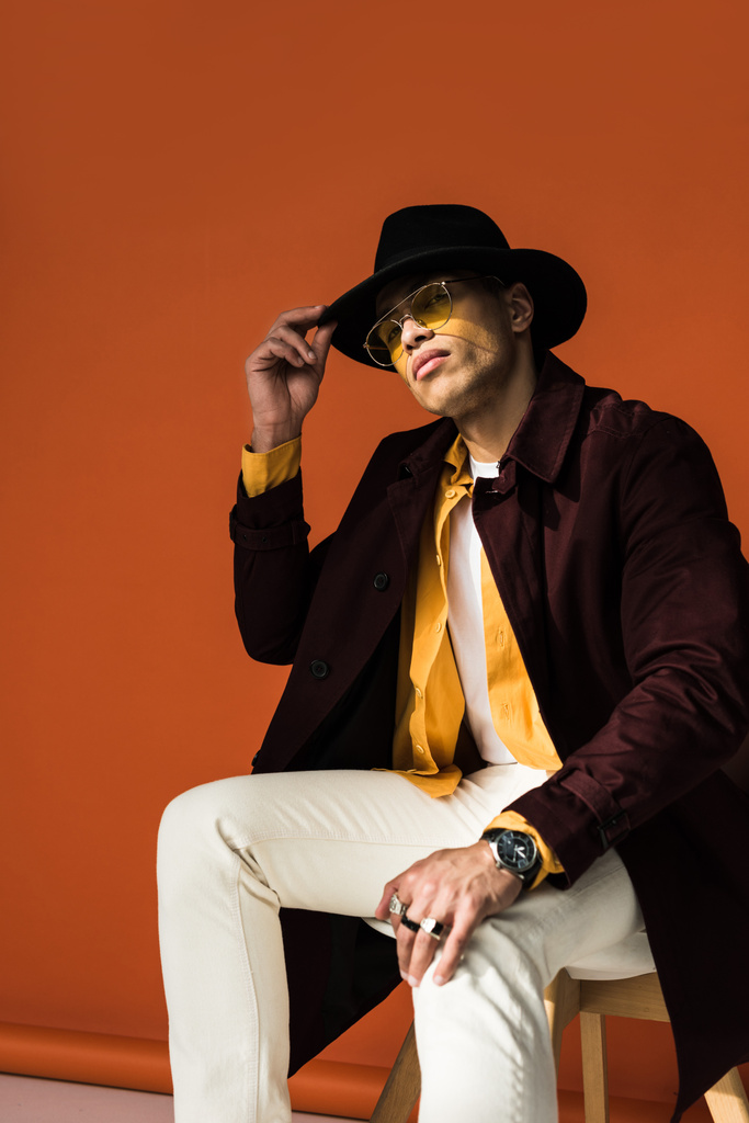 elegante uomo di razza mista in cappello e occhiali da sole seduto e in posa su arancione
 - Foto, immagini