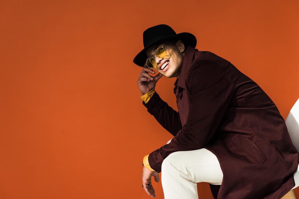 stylish mixed race man in hat and sunglasses sitting and looking at camera isolated on orange with copy space - Photo, Image