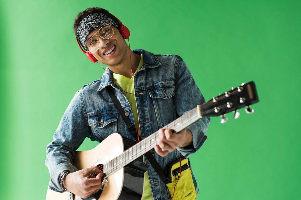 handsome mixed race man in denim and headphones looking at camera while playing acoustic guitar on green screen - Photo, Image