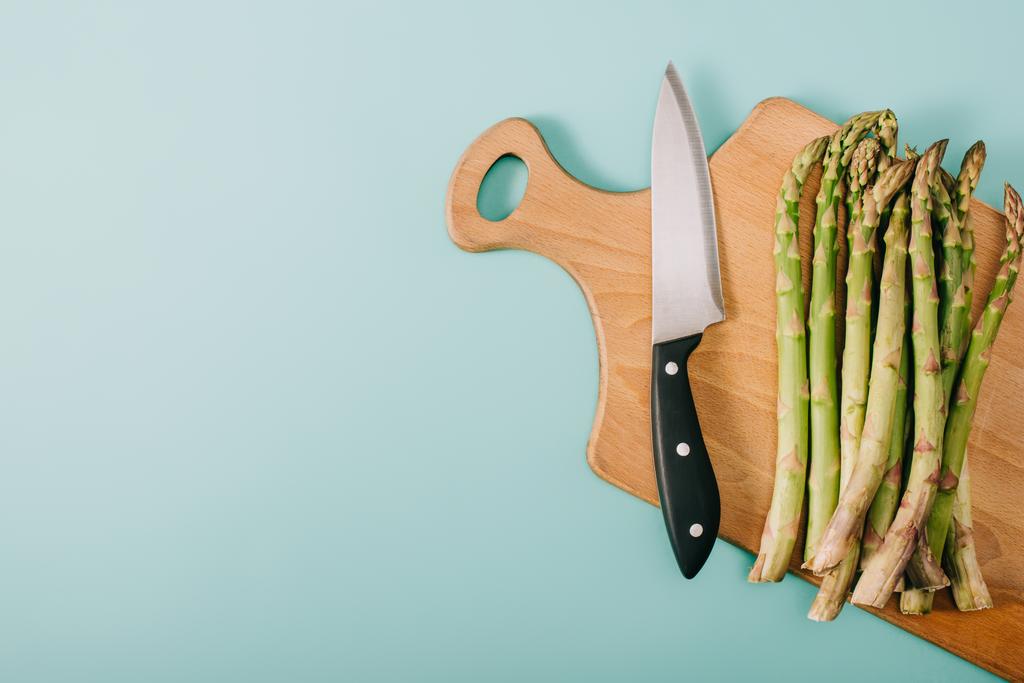 Ansicht von grünem rohen Spargel auf Holzschneidebrett mit Messer auf blauem Hintergrund - Foto, Bild