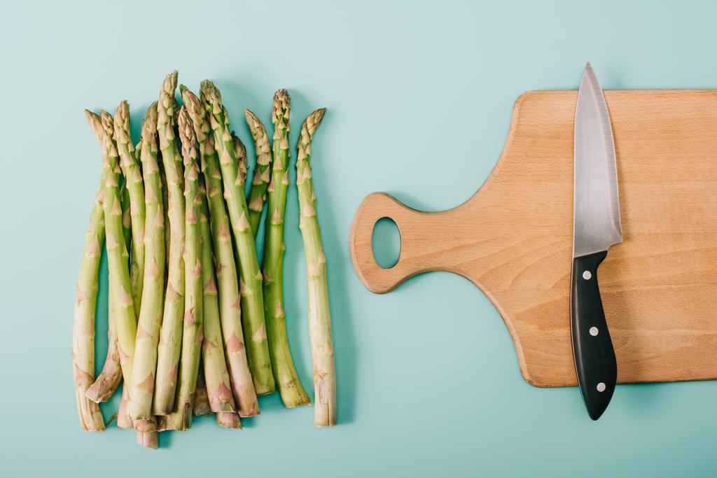 vista dall'alto di asparagi grezzi verdi vicino al tagliere in legno con coltello su sfondo blu
 - Foto, immagini