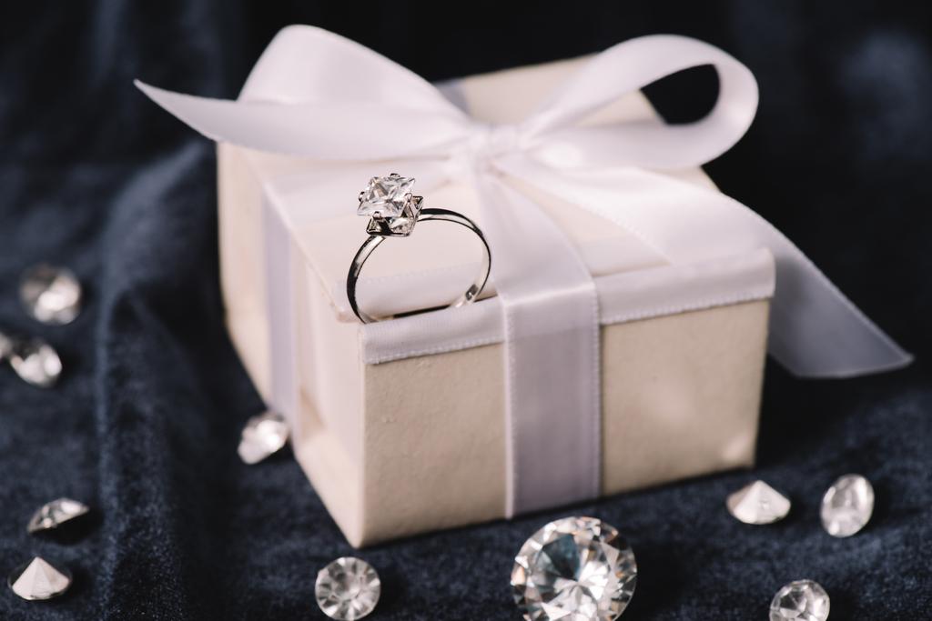 selective focus of engagement ring on gift box with bow near shiny diamonds on blue cloth - Photo, Image