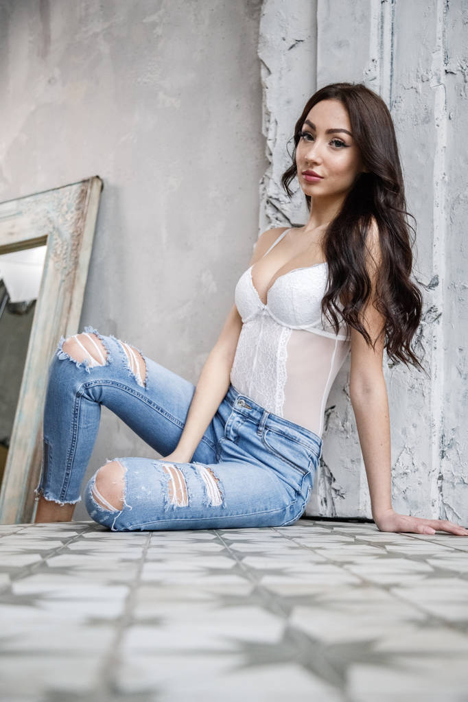 Studio portrait of beautiful brunette woman wearing white lingerie and blue jeans. Charming beauty model posing at apartments interior   - Photo, Image