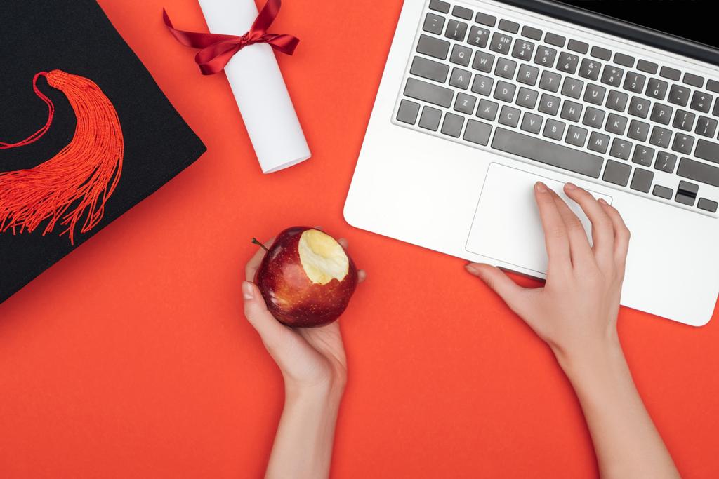 Vista cortada da menina com boné acadêmico, diploma e maçã usando laptop na superfície vermelha
 - Foto, Imagem
