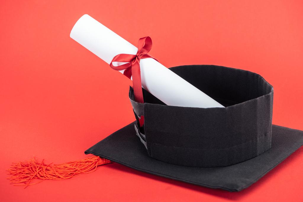 Academische cap met kwast en diploma met lint op rode oppervlak - Foto, afbeelding