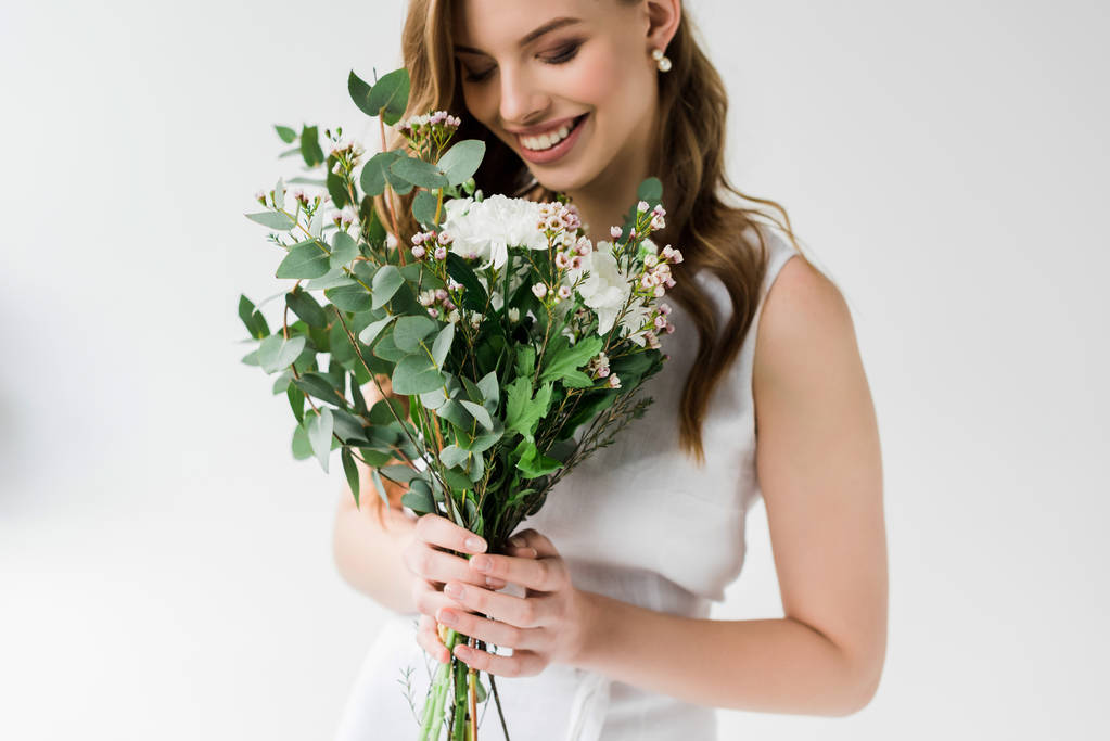 εύθυμη νεαρή γυναίκα κοιτάζοντας τα λουλούδια και χαμογελώντας στο λευκό - Φωτογραφία, εικόνα