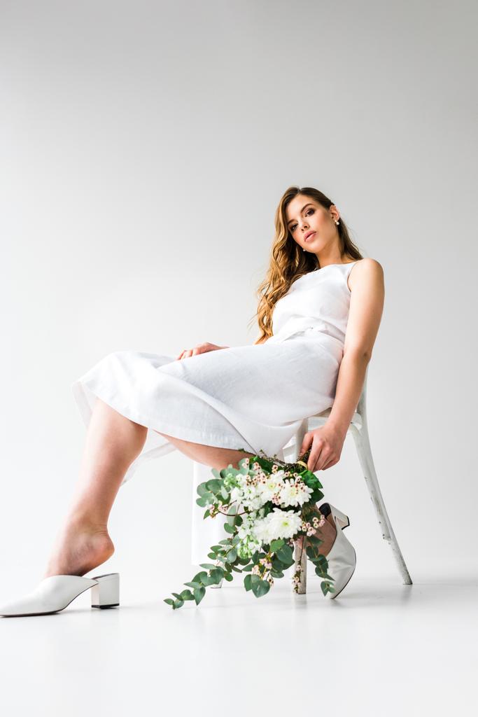 низкий угол зрения молодой женщины в платье сидя на стуле и держа букет цветов с эвкалиптовыми листьями на белом
  - Фото, изображение