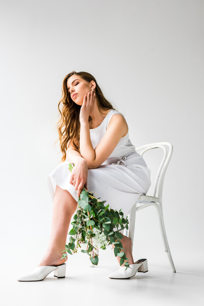 Nuori nainen mekko istuu tuolilla suljetuin silmin ja tilalla kukkakimppu eukalyptus lehdet valkoinen
  - Valokuva, kuva